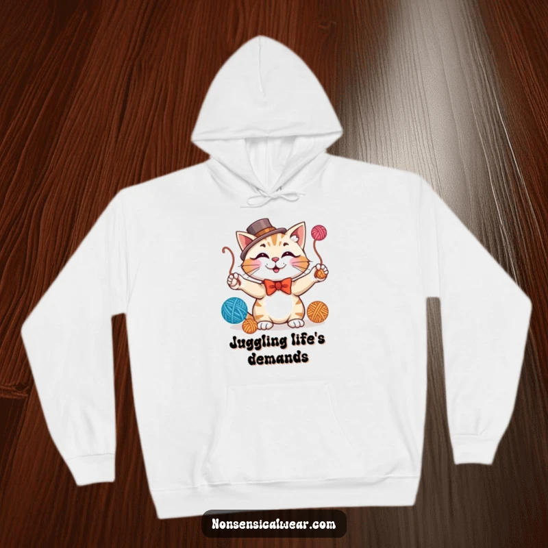 Funny hoodie depicting a smiling cat in a bowtie and top hat, expertly juggling balls of yarn, exuding charm.